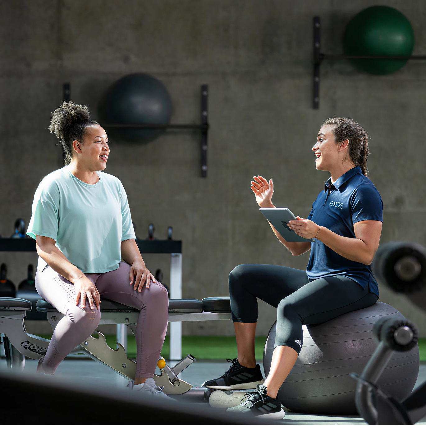 EXOS personal trainer consulting with a client in a modern gym setting, using tablet technology for personalized fitness coaching